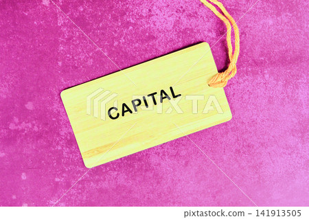 A tag with the word capital is on a pink surface. The tag has a natural wood look and a string attached. It seems to be part of a game or an activity A tag with the word capital is on a pink surface. The tag has a natural wood look and a string attached. It seems to be part of a game or an activity 141913505