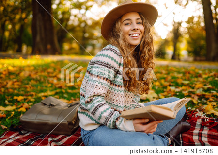 Tourist is reading a book and enjoying the autumn landscape in the park while sitting on plaid. 141913708