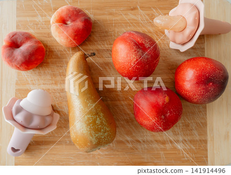Flat lay composition of whole ripe peaches, a pear, and silicone baby fruit feeders on a wooden cutting board. Perfect background for baby food recipes, teething, and healthy nutrition concepts Flat lay composition of whole ripe peaches, a pear, and silicone baby fruit feeders on a wooden cutting board. Perfect background for baby food recipes, teething, and healthy nutrition concepts 141914496