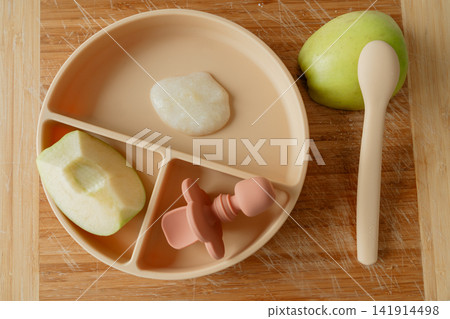 Top view of a neutral beige silicone baby plate with apple sauce, fresh green apple slice, spoon, and feeder on wooden board. Ideal for minimalist baby care, healthy food, and weaning stock themes Top view of a neutral beige silicone baby plate with apple sauce, fresh green apple slice, spoon, and feeder on wooden board. Ideal for minimalist baby care, healthy food, and weaning stock themes 141914498