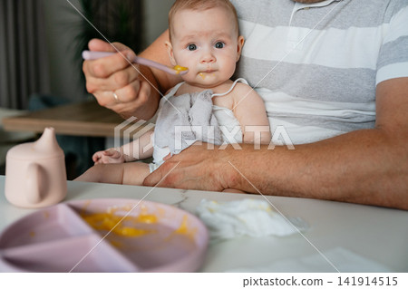 a baby being fed in a calm indoor home environment, with a high chair and spoon, viewed from the baby's perspective, wearing pink and white clothing, surrounded by a wooden high chair, evoc. a baby being fed in a calm indoor home environment, with a high chair and spoon, viewed from the baby's perspective, wearing pink and white clothing, surrounded by a wooden high chair, evoc. 141914515