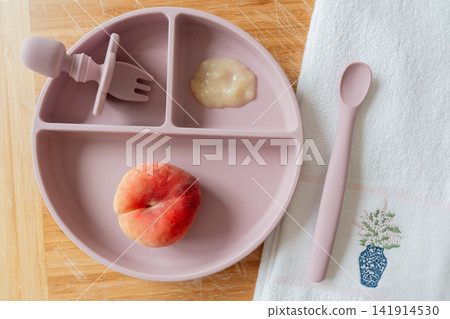 Baby meal setup feeding, in table, with baby plate, fork, spoon, peach, from a top-down perspective, with pink, white accents, and calm mood, showing nearby objects and surrounding setting. Baby meal setup feeding, in table, with baby plate, fork, spoon, peach, from a top-down perspective, with pink, white accents, and calm mood, showing nearby objects and surrounding setting. 141914530