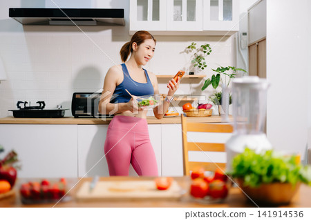 Fitness woman reading recipe while preparing healthy smoothie with fresh vegetables in modern kitchen. Healthy lifestyle, nutrition, home cooking Fitness woman reading recipe while preparing healthy smoothie with fresh vegetables in modern kitchen. Healthy lifestyle, nutrition, home cooking 141914536
