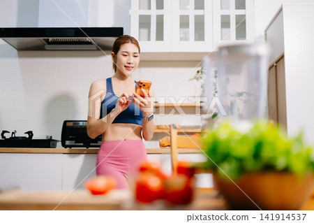 Fitness woman reading recipe while preparing healthy smoothie with fresh vegetables in modern kitchen. Healthy lifestyle, nutrition, home cooking 141914537