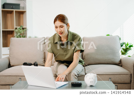 Smart financial management. Young Asian woman planning budget and savings using laptop, calculator, and piggy bank in a bright living room 141914548