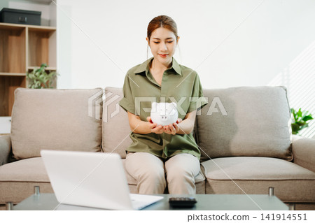 Smart financial management. Young Asian woman planning budget and savings using laptop, calculator, and piggy bank in a bright living room 141914551