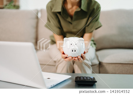 Smart financial management. Young Asian woman planning budget and savings using laptop, calculator, and piggy bank in a bright living room Smart financial management. Young Asian woman planning budget and savings using laptop, calculator, and piggy bank in a bright living room 141914552
