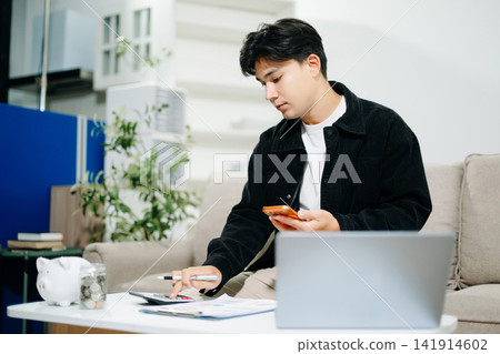 Young man using a tablet for budgeting and financial planning at home. Savings, fintech, money management, investment goals and digital finance concept. Young man using a tablet for budgeting and financial planning at home. Savings, fintech, money management, investment goals and digital finance concept. 141914602