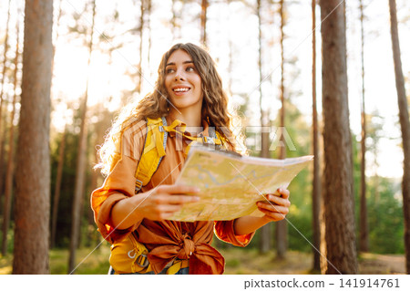 A traveler woman stands in a lush forest, examining a map with a focused expression A traveler woman stands in a lush forest, examining a map with a focused expression 141914761