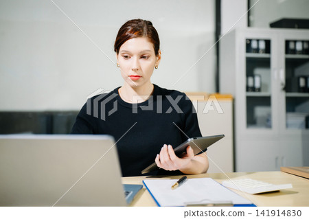 Young woman typing on tablet and laptop while sitting at the working white table in modern office Young woman typing on tablet and laptop while sitting at the working white table in modern office 141914830