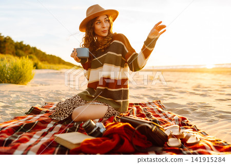 Happy woman in a stylish sweater sits at picnic on the beach drinks a hot drink from a thermos Happy woman in a stylish sweater sits at picnic on the beach drinks a hot drink from a thermos 141915284
