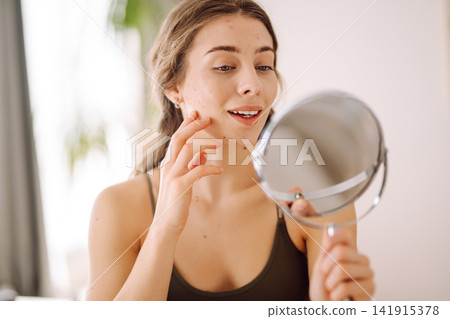Young woman with problem skin looking into mirror. Inflammation on face, problem skin. Beauty care. Young woman with problem skin looking into mirror. Inflammation on face, problem skin. Beauty care. 141915378