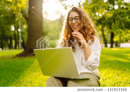 Young woman in glasses and with laptop working at sunset in park on green meadow. Lifestyle concept. Young woman in glasses and with laptop working at sunset in park on green meadow. Lifestyle concept. 141915568
