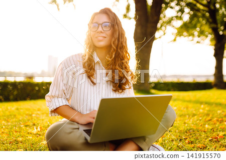Young Woman sitting on grass ground, working on laptop. Freelance work, technology concept. Young Woman sitting on grass ground, working on laptop. Freelance work, technology concept. 141915570