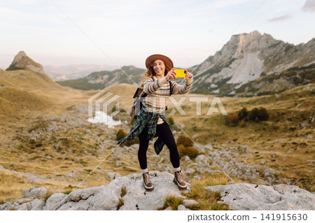 Woman stands on rocky terrain, phone in hand, taking picture of breathtaking landscape. Blogging. Woman stands on rocky terrain, phone in hand, taking picture of breathtaking landscape. Blogging. 141915630