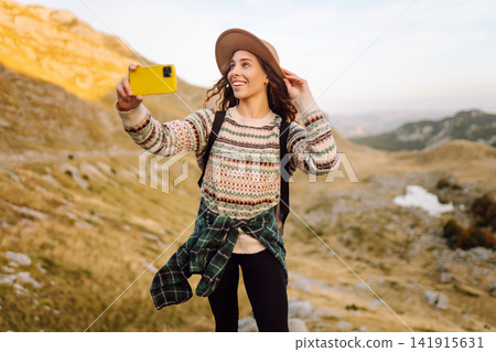 Female traveler with single backpack takes selfie against backdrop of mountain scenery. Lifestyle. Female traveler with single backpack takes selfie against backdrop of mountain scenery. Lifestyle. 141915631