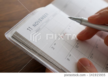 特寫鏡頭:一位女士在木桌旁,正在十一月的日程表上寫字,鏡頭對準她的雙手。 特寫鏡頭:一位女士在木桌旁,正在十一月的日程表上寫字,鏡頭對準她的雙手。 141915935
