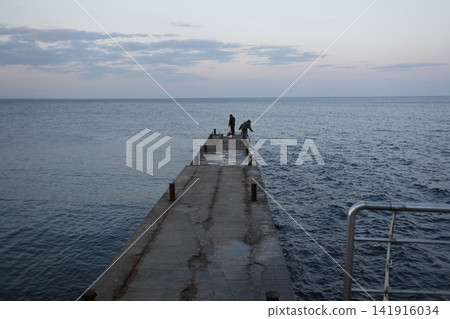 Long concrete pier extending into calm open sea beneath a cloudy evening sky, with distant people at the end, capturing coastal tranquility, perspective and peaceful marine scenery Long concrete pier extending into calm open sea beneath a cloudy evening sky, with distant people at the end, capturing coastal tranquility, perspective and peaceful marine scenery 141916034