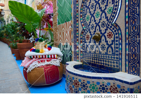 Colorful Moroccan tiles frame a decorative brass fountain beside stacked cushions, textiles and potted plants in a narrow medina alley, creating a cozy, vibrant traditional North African scene. Colorful Moroccan tiles frame a decorative brass fountain beside stacked cushions, textiles and potted plants in a narrow medina alley, creating a cozy, vibrant traditional North African scene. 141916511