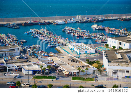 Aerial view of a busy fishing harbor filled with colorful boats, piers and a seaside market along turquoise Mediterranean waters, showing maritime industry, coastal life and local activity. Aerial view of a busy fishing harbor filled with colorful boats, piers and a seaside market along turquoise Mediterranean waters, showing maritime industry, coastal life and local activity. 141916532