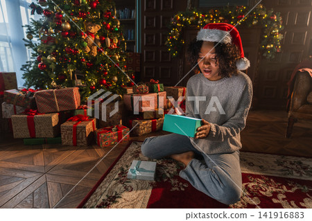 Merry Christmas. African American woman unpacking open up gift box near Christmas tree. Girl in living room with Christmas tree and fireplace opening gift box with surprise face. Christmas eve at home Merry Christmas. African American woman unpacking open up gift box near Christmas tree. Girl in living room with Christmas tree and fireplace opening gift box with surprise face. Christmas eve at home 141916883
