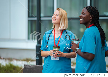 Diverse female medical workers smiling having coffee break outdoors Diverse female medical workers smiling having coffee break outdoors 141917191