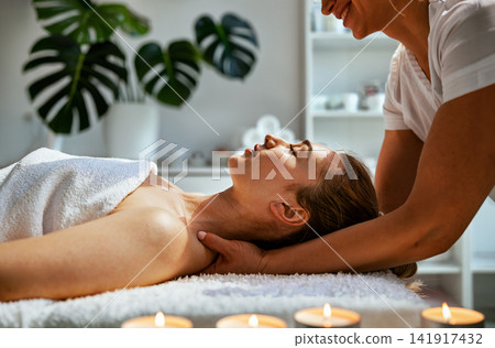 Neck and Decolletage Massage at Biophilic Wellness Spa Smiling Therapist Lifting Womans Shoulders by Candlelight with Monstera Plants Behind Neck and Decolletage Massage at Biophilic Wellness Spa Smiling Therapist Lifting Womans Shoulders by Candlelight with Monstera Plants Behind 141917432