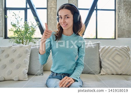 Young woman in headphones showing thumbs up gesture sitting on sofa Young woman in headphones showing thumbs up gesture sitting on sofa 141918019