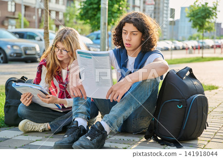 Two teenage students sitting outdoors reading workbook preparing for exams together Two teenage students sitting outdoors reading workbook preparing for exams together 141918044