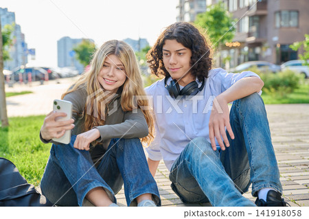 Happy teenage couple taking selfie with smartphone outdoors sitting on ground Happy teenage couple taking selfie with smartphone outdoors sitting on ground 141918058