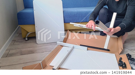 Person assembling white furniture from flat pack box on wooden floor Person assembling white furniture from flat pack box on wooden floor 141919844