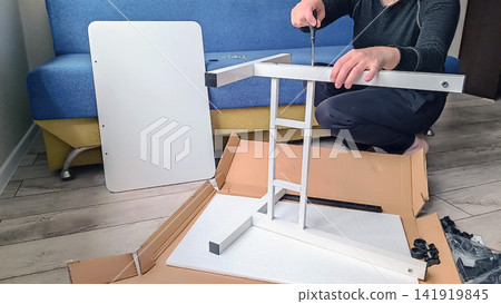 Person assembling white furniture frame with screwdriver on wooden floor Person assembling white furniture frame with screwdriver on wooden floor 141919845