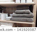 A neat stack of four grey terry towels alongside white embossed ceramic tumblers and a soap dish on light wood retail shelves, displaying minimalist bathroom accessories. 141920387