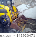 A close-up view of a worn, yellow skid-steer loader with its rusty metal front bucket resting on a dirty ground surface next to a peeling, weathered concrete wall. 141920471