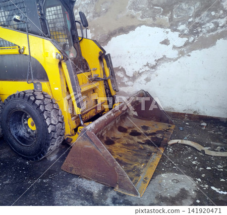 A close-up view of a worn, yellow skid-steer loader with its rusty metal front bucket resting on a dirty ground surface next to a peeling, weathered concrete wall. A close-up view of a worn, yellow skid-steer loader with its rusty metal front bucket resting on a dirty ground surface next to a peeling, weathered concrete wall. 141920471