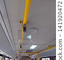An interior perspective looking up at the ceiling of a bus. A clear plastic grab handle is hanging from a yellow horizontal handrail via a grey strap. Background shows lights and more railings. 141920472