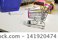 Miniature chrome-wire shopping cart with pink plastic accents sits on a white display table, casting a shadow. Blue crate and store items are in the background. 141920474