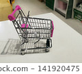 A miniature metal shopping cart with bright pink plastic handle and seat accents sits empty on a white surface in a store. It casts a shadow. Blurred shop shelves are in the background. 141920475