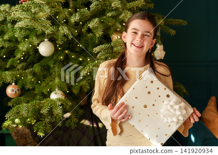 Teen girl holding Christmas gifts and smiling by festive tree Teen girl holding Christmas gifts and smiling by festive tree 141920934
