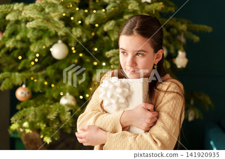 Teen girl hugging Christmas present beside decorated holiday tree Teen girl hugging Christmas present beside decorated holiday tree 141920935