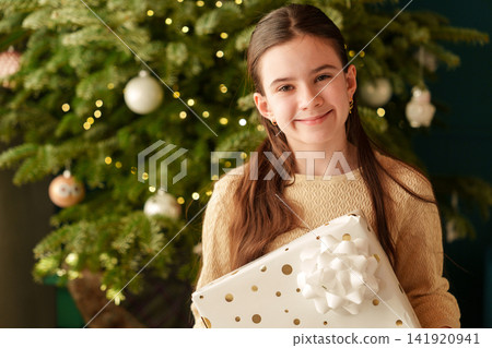 Teen girl holding large Christmas gift by decorated tree at home 141920941
