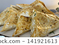 A large plate holds many pieces of pastry that are cut into triangles. They are golden brown and placed on a white dish at an event. A person stands nearby, serving guests. 141921613