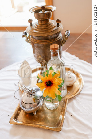 Close-up photo of an antique brass Russian samovar on a tray with a small vase holding a yellow sunflower. Ideal for Russian tea party tradition, folk culture, and vintage heritage concepts Close-up photo of an antique brass Russian samovar on a tray with a small vase holding a yellow sunflower. Ideal for Russian tea party tradition, folk culture, and vintage heritage concepts 141922127