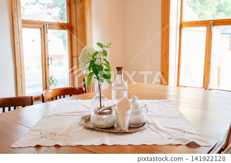 Sunlit interior of a countryside house dining table with glass bottle, sugar bowl, and white hydrangeas. Perfect stock photo for eco-friendly living, family home, and peaceful mornings Sunlit interior of a countryside house dining table with glass bottle, sugar bowl, and white hydrangeas. Perfect stock photo for eco-friendly living, family home, and peaceful mornings 141922128
