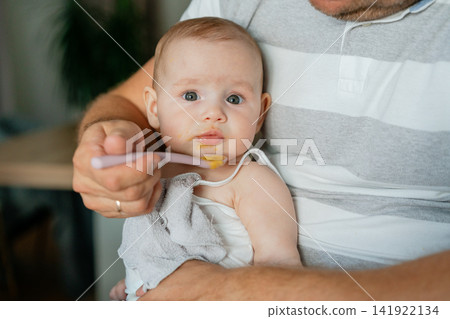 Portrait of a cute baby sitting in father's arms looking at camera while being spoon-fed. Perfect for pediatric care, baby food marketing, and family lifestyle promotional materials Portrait of a cute baby sitting in father's arms looking at camera while being spoon-fed. Perfect for pediatric care, baby food marketing, and family lifestyle promotional materials 141922134