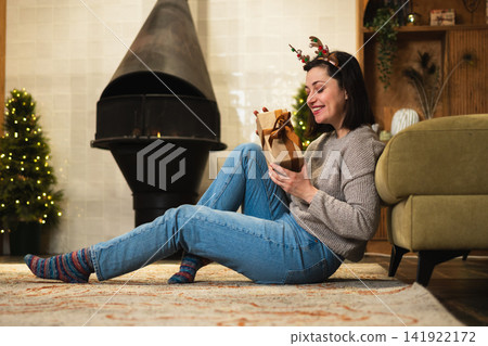 Merry Christmas. Young adult woman in unpacking open up gift box near Christmas tree. Girl in living room with Christmas tree, fireplace opening gift box with surprise face. Christmas eve at home 141922172