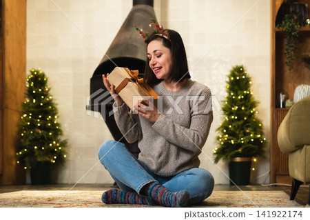 Merry Christmas. Young adult woman in unpacking open up gift box near Christmas tree. Girl in living room with Christmas tree, fireplace opening gift box with surprise face. Christmas eve at home Merry Christmas. Young adult woman in unpacking open up gift box near Christmas tree. Girl in living room with Christmas tree, fireplace opening gift box with surprise face. Christmas eve at home 141922174