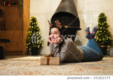 Merry Christmas. Young adult woman in unpacking open up gift box near Christmas tree. Girl in living room with Christmas tree, fireplace opening gift box with surprise face. Christmas eve at home 141922175