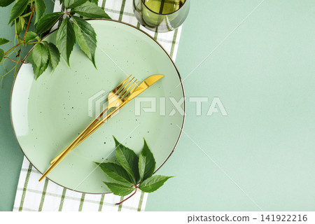 Elegant table setting with pastel plate golden cutlery green leaves and checkered napkin for seasonal celebration Elegant table setting with pastel plate golden cutlery green leaves and checkered napkin for seasonal celebration 141922216