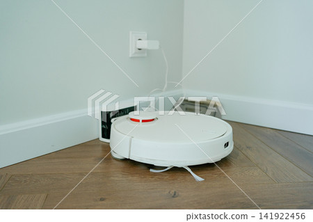 Robotic vacuum cleaner charging near a light wall on hard wood floor. Perfect stock photo for smart home technology, daily household chores, modern domestic gadgets, and interior cleanliness Robotic vacuum cleaner charging near a light wall on hard wood floor. Perfect stock photo for smart home technology, daily household chores, modern domestic gadgets, and interior cleanliness 141922456